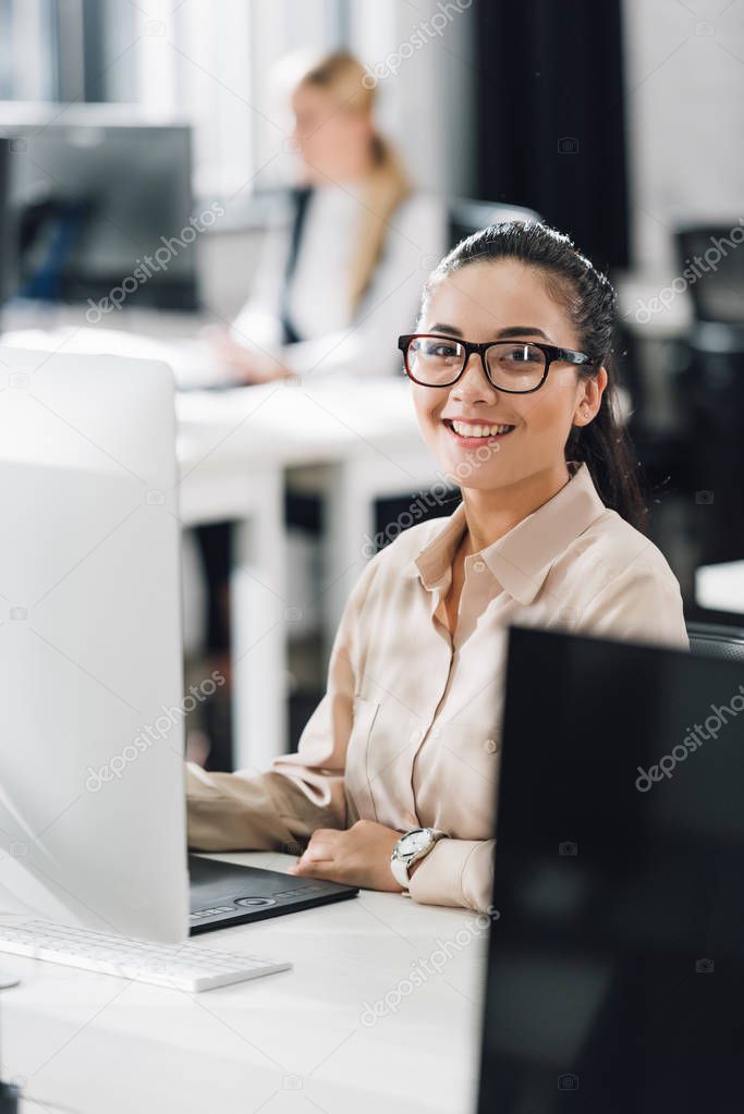 attractive young businesswoman using desktop computer and smiling at camera in office 