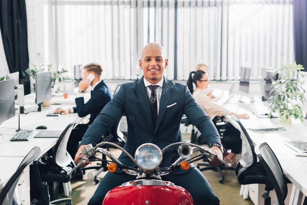 happy young african american businessman riding red motorbike and smiling at camera in office  
