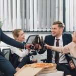 Jovens colegas felizes bebendo cerveja e comendo pizza no escritório