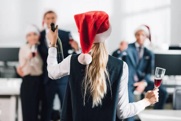 在与同事一起庆祝圣诞节的同时 穿着圣诞老人帽拿着玻璃和香槟酒的女商人的背影 — 图库照片