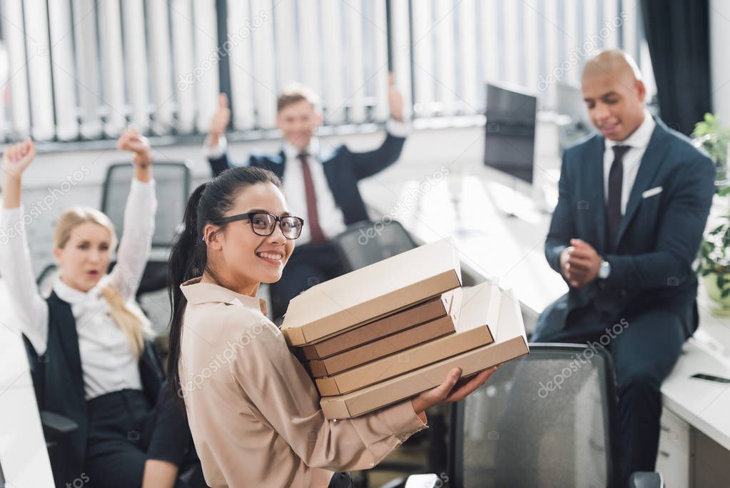 young businesswoman holding pizza boxes and smiling at camera while coworkers sitting behind in open space office