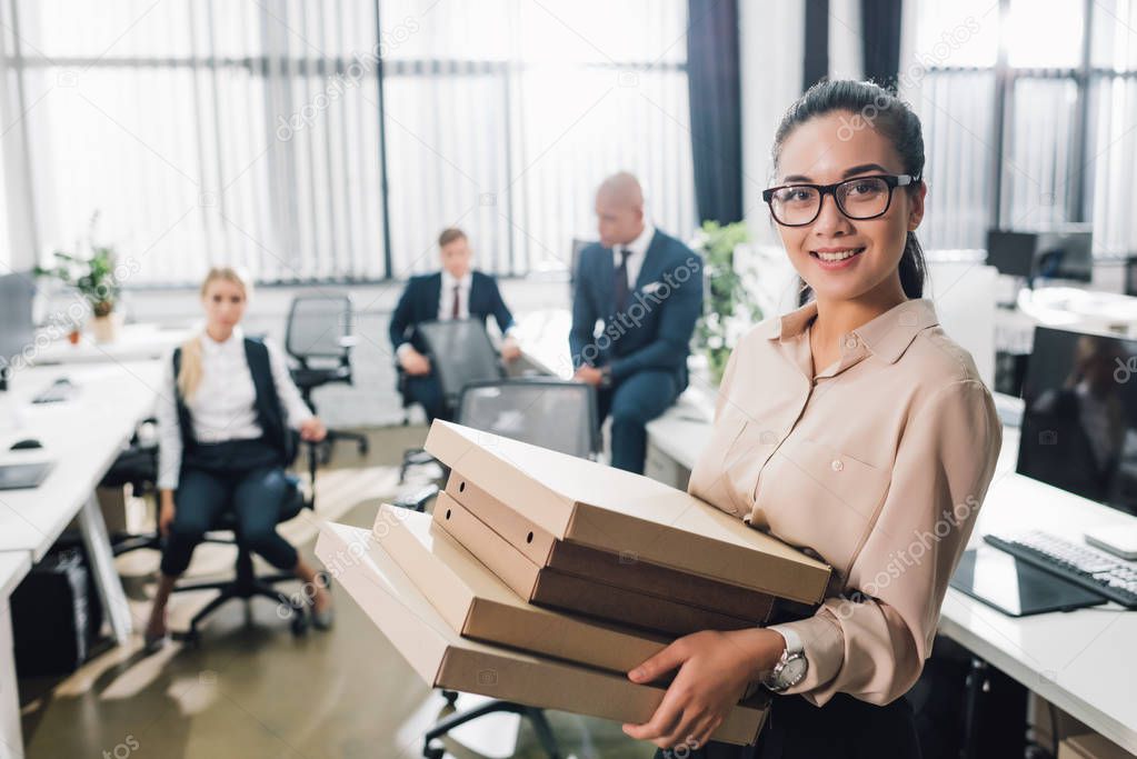 young businesswoman holding pizza boxes and smiling at camera while colleagues sitting behind in office  