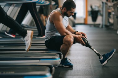 yorgun genç sporcu spor salonunda koşu bandı üzerinde oturan yapay bacak ile yan görünüm