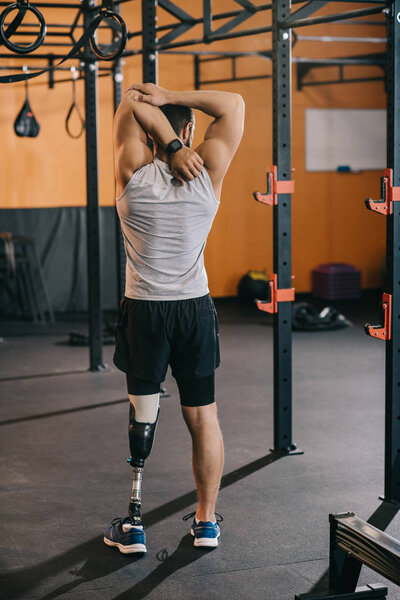 вид сзади на молодого спортсмена с искусственной ногой, растянувшейся возле гимнастической лестницы в спортзале
