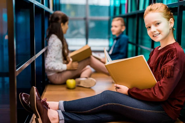 adorable schoolchild holding book and smiling at camera while studying with friends in library