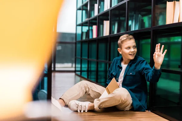 Leuke Jongen Bedrijf Boek Glimlachen Zwaaien Hand Zittend Vloer Bibliotheek — Gratis stockfoto