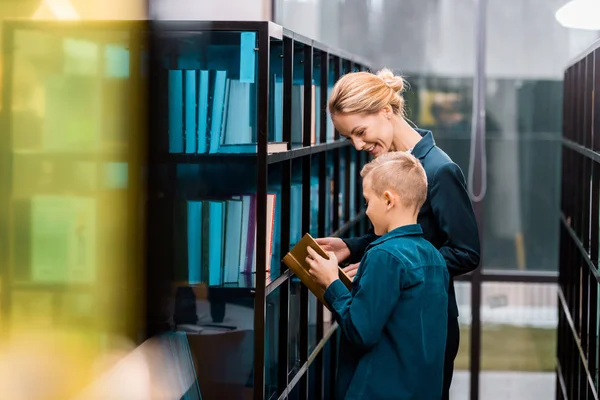 Uśmiechnięta Młoda Kobieta Bibliotekarz Ogląda Uczniak Gospodarstwa Książki Bibliotece — Zdjęcie stockowe