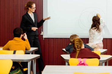 young teacher showing the whiteboard and schoolkid writing during presentation clipart