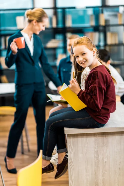 Adorable Schoolkid Holding Book Smiling Camera While Visiting Library Classmates — Stock Photo, Image