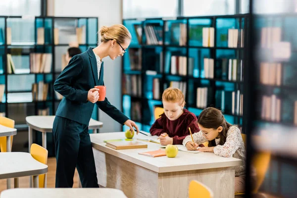 Joven Profesora Sosteniendo Taza Mirando Las Colegialas Escribiendo Estudiando Biblioteca — Foto de Stock