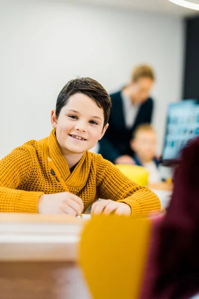 Bedårande Skolpojke Skriver Med Penna Och Ler Mot Kameran — Gratis stockfoto