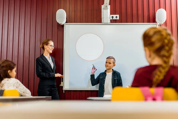 teacher and schoolchildren looking at boy making presentation at interactive whiteboard