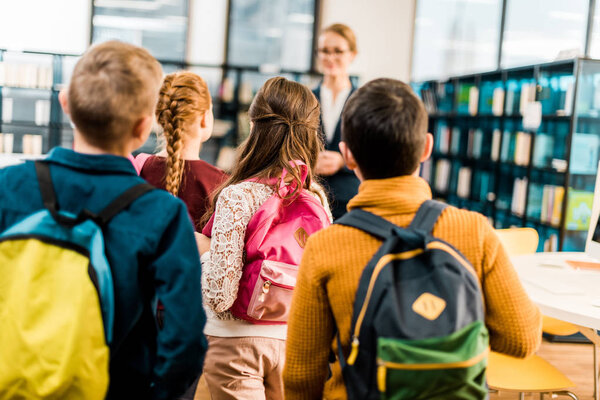 back view of schoolchildren with backpacks looking at librarian in library 