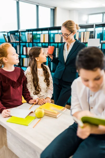 Smilende Ung Kvinnelig Lærer Som Holder Kopp Ser Skolebarn Biblioteket stockbilde