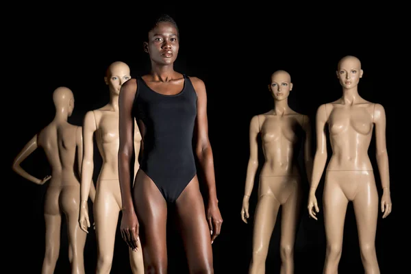 Красивая африканская американка в боди-костюме, стоящая перед манекенами и смотрящая в камеру на черном — стоковое фото