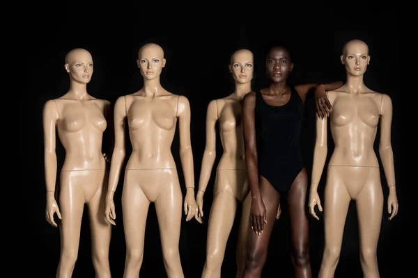 Beautiful young african american woman in swimsuit standing between mannequins and looking at camera on black — Stock Photo