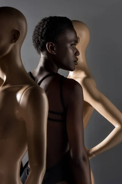 Вид збоку красивої афроамериканської дівчини, яка дивиться далеко, стоячи між манекенами на чорному — стокове фото