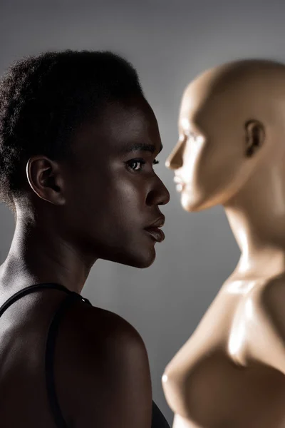 Profile portrait of beautiful african american girl looking away while standing near mannequin on black — Stock Photo