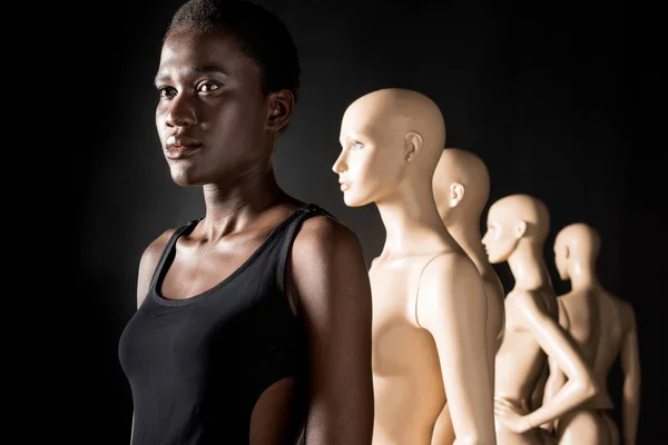 Afroamericana chica de pie en fila con maniquíes y mirando hacia otro lado en negro - foto de stock
