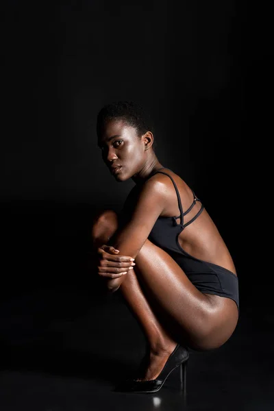 Vista lateral de la hermosa chica afroamericana en traje de cuerpo agachado y mirando a la cámara en negro - foto de stock