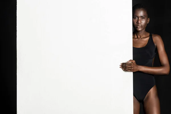 Африканская американка в боди-костюме позирует с пустой белой картой и смотрит в камеру на черном — стоковое фото