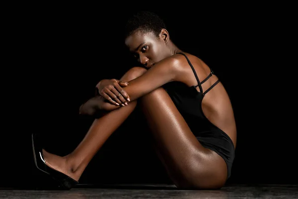 Вид сбоку привлекательной африканской девушки в купальнике, сидящей и смотрящей на камеру в черном — стоковое фото