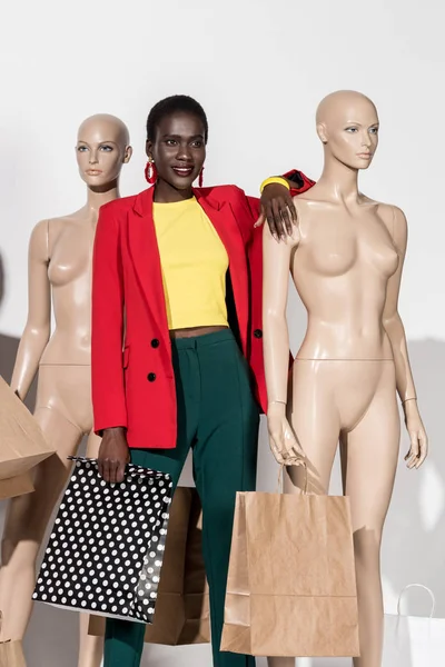 Красивая улыбающаяся африканская американка, стоящая между манекенами с пакетами покупок на белом — стоковое фото