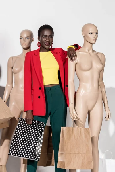 Hermosa sonriente afroamericana chica de pie entre maniquíes con bolsas de compras en blanco - foto de stock