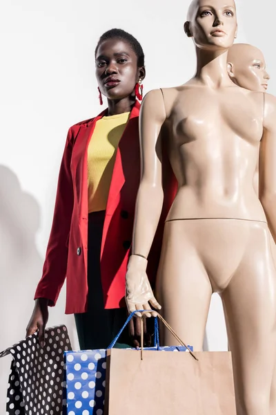 Vue à angle bas de la jeune femme afro-américaine regardant la caméra tout en se tenant près des mannequins avec des sacs à provisions sur blanc — Photo de stock