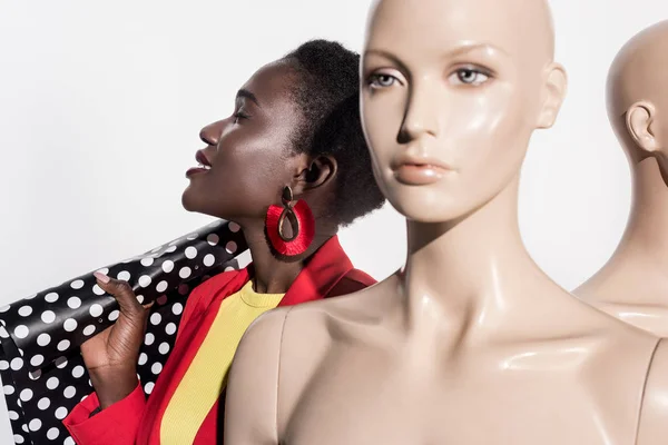 Smiling young african american woman holding shopping bag and standing near mannequins on white — Stock Photo