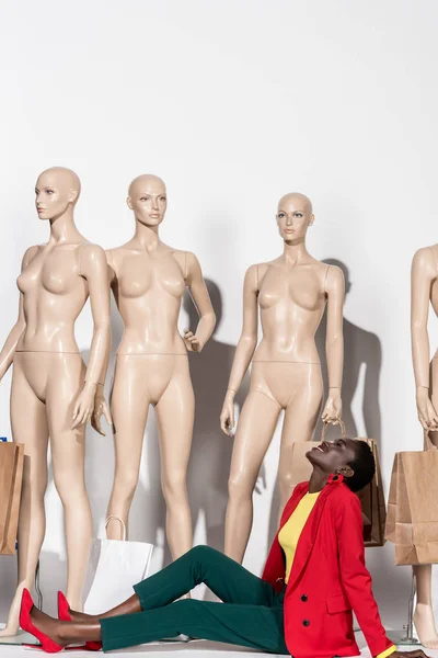 Sorrindo mulher americana africana elegante olhando para cima enquanto sentado na frente de manequins com sacos de compras — Fotografia de Stock