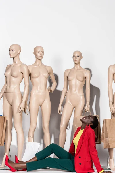 Smiling stylish african american woman looking up while sitting in front of mannequins with shopping bags — Stock Photo