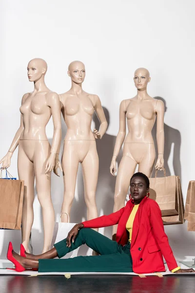 Mujer afroamericana de moda mirando a la cámara mientras está sentado frente a maniquíes con bolsas de compras - foto de stock