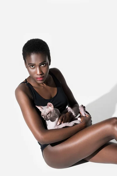 Vista de ángulo alto de la hermosa chica afroamericana en traje de cuerpo sentado con gato y mirando a la cámara en blanco - foto de stock