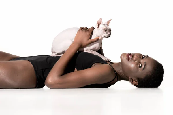 Vista lateral de mujer afroamericana en traje de baño acostado con gato y mirando a la cámara aislada en blanco - foto de stock