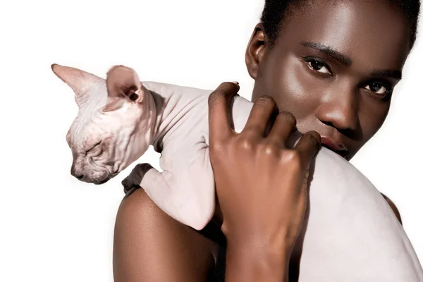 Vista de cerca de la hermosa chica afroamericana sosteniendo gato esfinge y mirando a la cámara aislada en blanco - foto de stock
