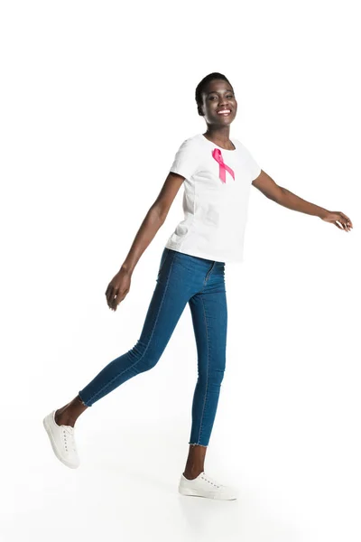 Allegra donna afroamericana con nastro rosa sulla t-shirt che cammina e sorride alla telecamera isolata sul bianco, concetto di cancro al seno — Foto stock