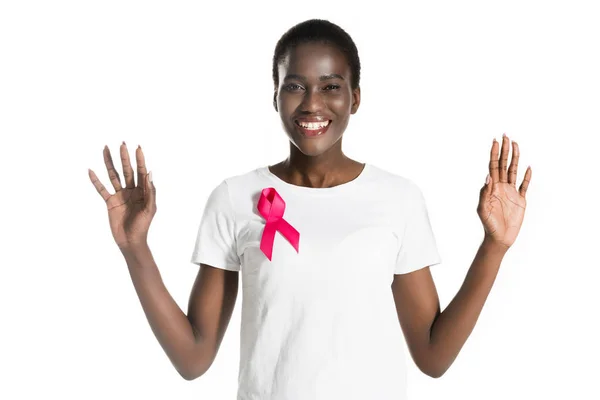 Cheerful african american girl with pink ribbon on t-shirt smiling at camera and raising hands isolated on white, breast cancer concept — Stock Photo