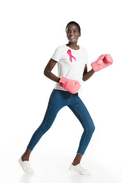 Beautiful african american girl with pink ribbon boxing and smiling at camera isolated on white, breast cancer concept — Stock Photo