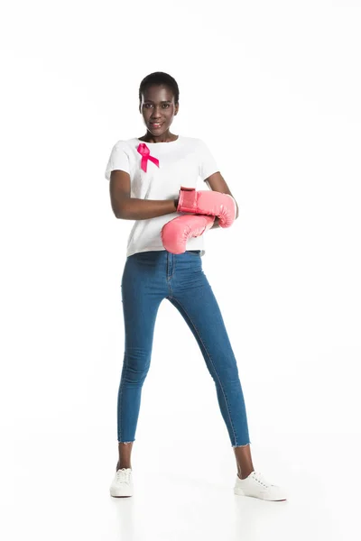 Vista completa de la mujer joven con cinta rosa con guantes de boxeo y sonriendo a la cámara aislada en blanco, concepto de cáncer de mama - foto de stock