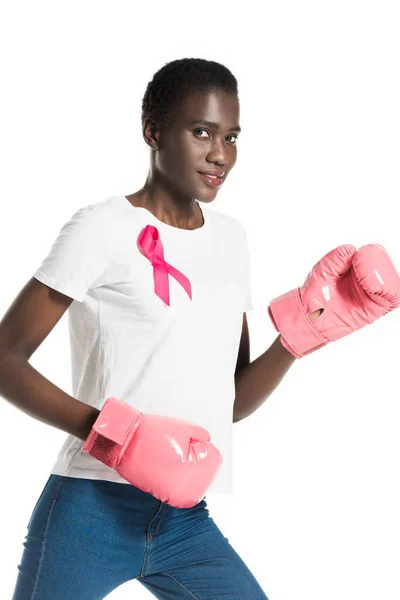 Вид сбоку на молодую женщину с розовой лентой на футболке боксера и улыбкой перед камерой, изолированной на белом, концепция рака груди — стоковое фото