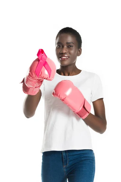 Jolie fille afro-américaine en gants de boxe tenant ruban rose et souriant à la caméra isolé sur blanc, concept de cancer du sein — Photo de stock