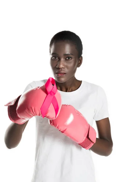 Young african american woman in boxing gloves holding pink ribbon and looking at camera isolated on white, breast cancer concept — Stock Photo