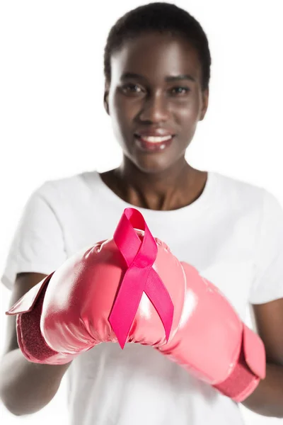 Close-up view of young african american woman in boxing gloves holding pink ribbon and smiling at camera isolated on white, breast cancer concept — Stock Photo