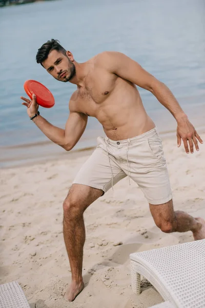 Schöner junger Mann mit fliegender Scheibe und Blick auf Kamera am Sandstrand — Stockfoto