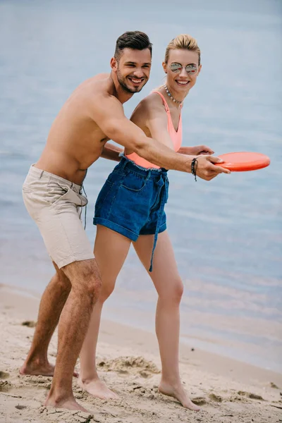 Feliz jovem casal brincando com disco voador na praia de areia — Fotografia de Stock