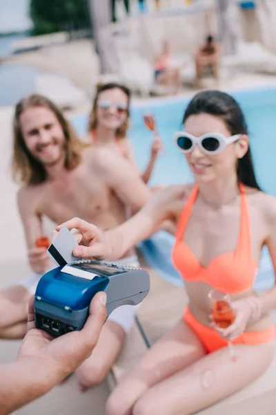Joven sonriente que paga con tarjeta de crédito mientras bebe champán con amigos en la piscina — Stock Photo