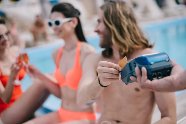 Lächelnder junger Mann, der mit Kreditkarte bezahlt und am Pool Champagner trinkende Freundinnen betrachtet — Stockfoto