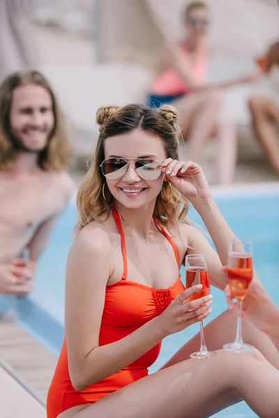 Счастливая молодая женщина в купальниках и солнцезащитных очках держит шампанское и улыбается перед камерой у бассейна — стоковое фото
