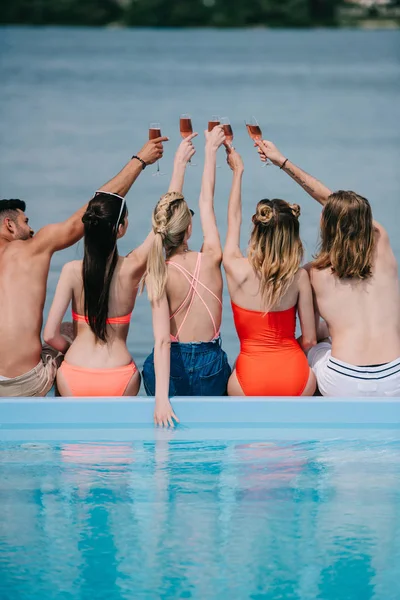 Visão traseira de homens e mulheres jovens que clinking copos de vinho enquanto sentado perto da piscina na praia — Fotografia de Stock